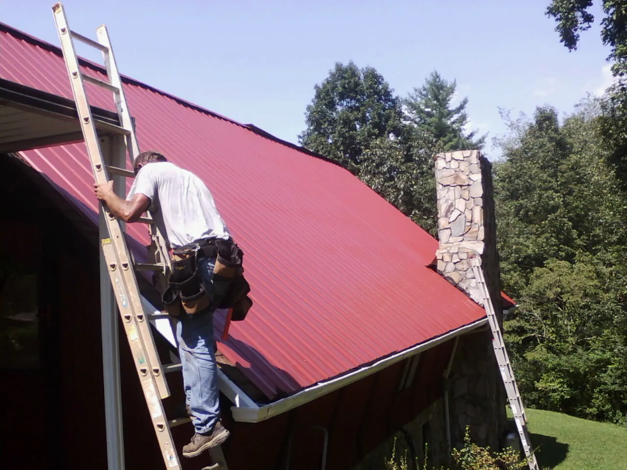 Roofer on a ladder beside a red metal roof and stone chimney — Soffit & Fascia Repair by Roofing Brothers in East Prospect